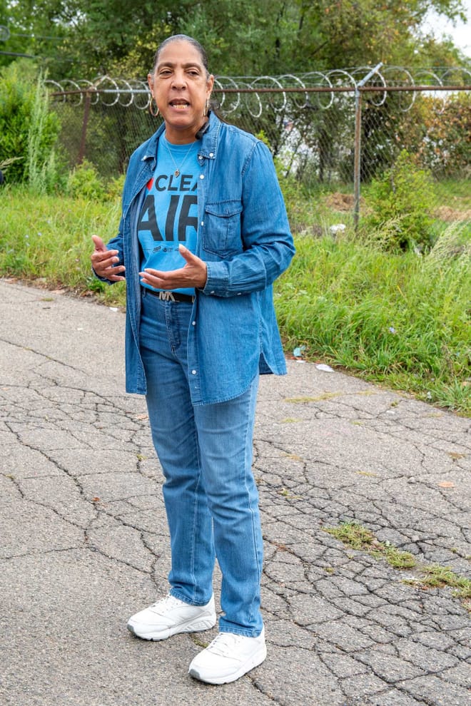 A woman with medium-tone skin and dark hair pulled back wearing jeans, a denim shirt and a blue t-shirt that says clean air