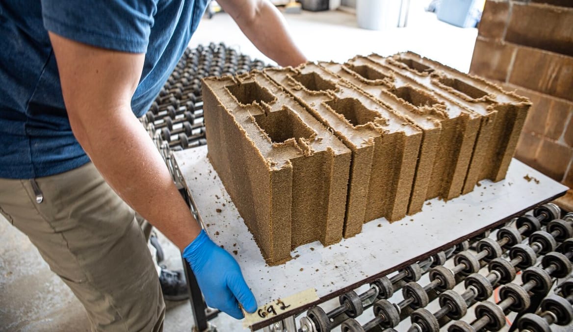 A human figure with khaki pants and blue latex gloves moves a tray of brown bricks