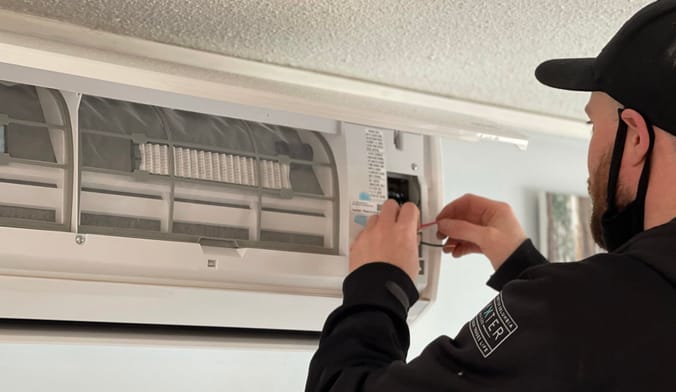 A technician wearing black clothing installs an indoor heat pump unit.