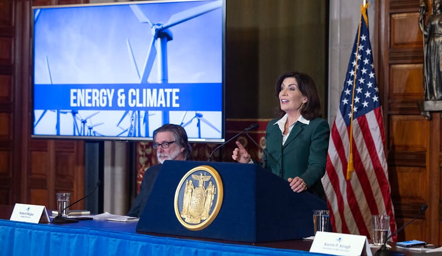 A woman in a green suit stands at a lectern with a golden state seal next to a video screen with the words energy and climate