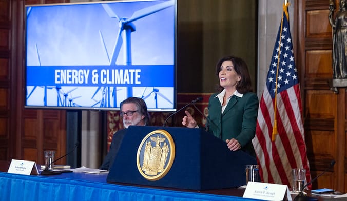 A woman in a green suit stands at a lectern with a golden state seal next to a video screen with the words energy and climate