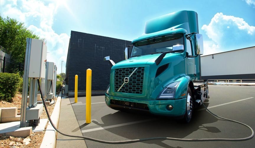 a teal semi truck attached to an EV charger in an industrial parking lot