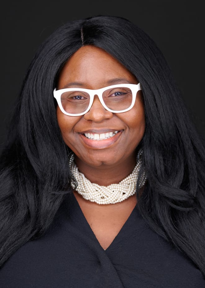 A smiling Black woman with long black hair, white glasses, a white beaded necklace and a black blouse