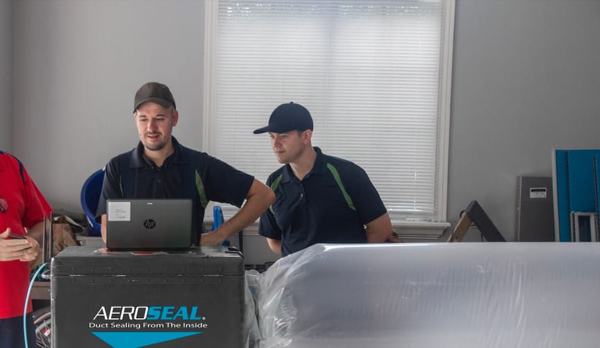 Two men in work uniforms look at a machine labeled 'Aeroseal' that is connected to a plastic tube