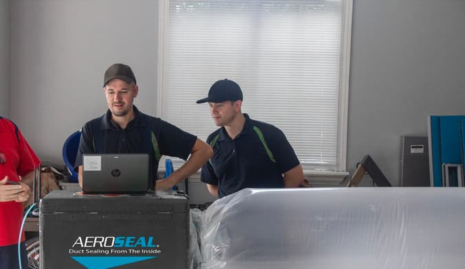 Two men in work uniforms look at a machine labeled 'Aeroseal' that is connected to a plastic tube