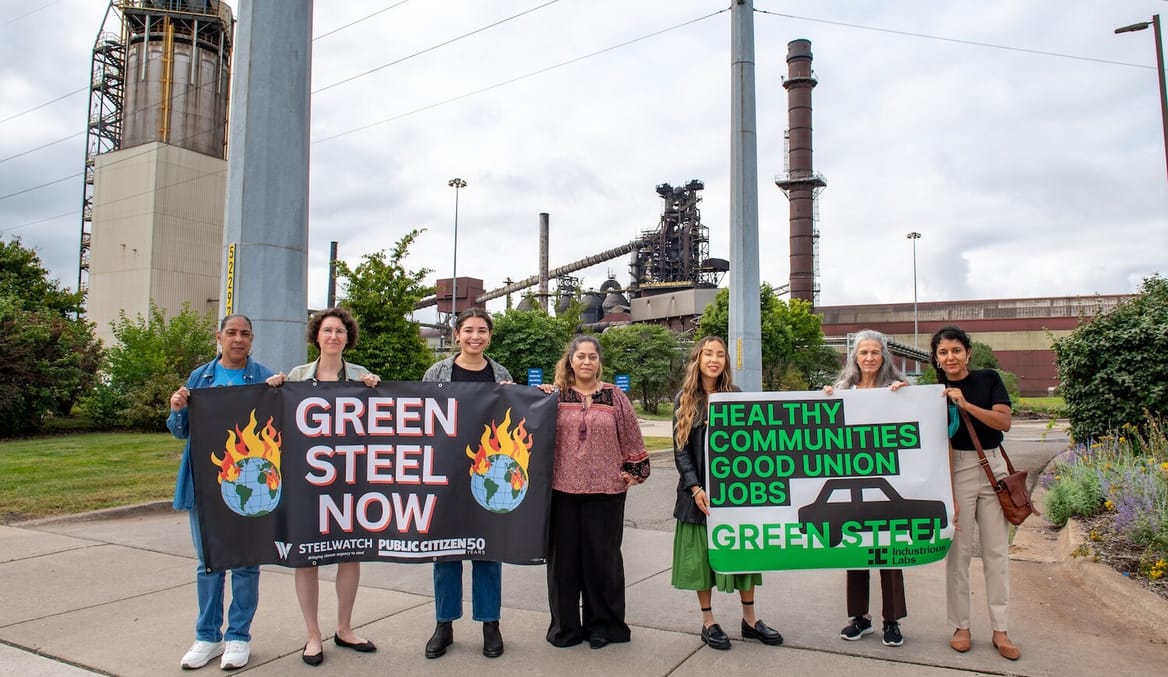 A group of women holding pro-green-steel signs stand in front of an industrial facility