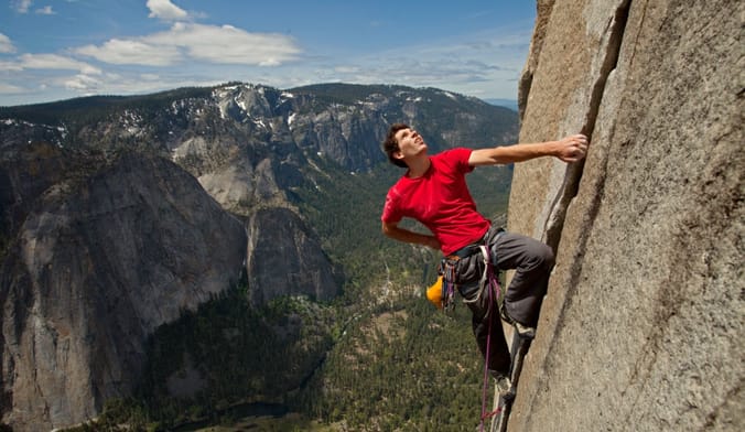 A man with a light skin tone wearing a red shirt and gray pants climbs a sheer rock face. A large mountain range looms behind