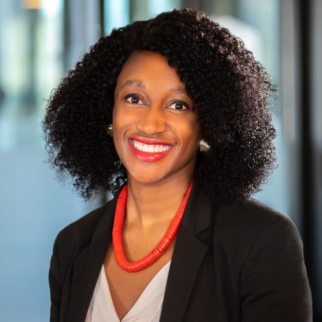 An African American woman with shoulder length dark brown hair, a black blazer, white v-neck blouse, and red-orange necklace