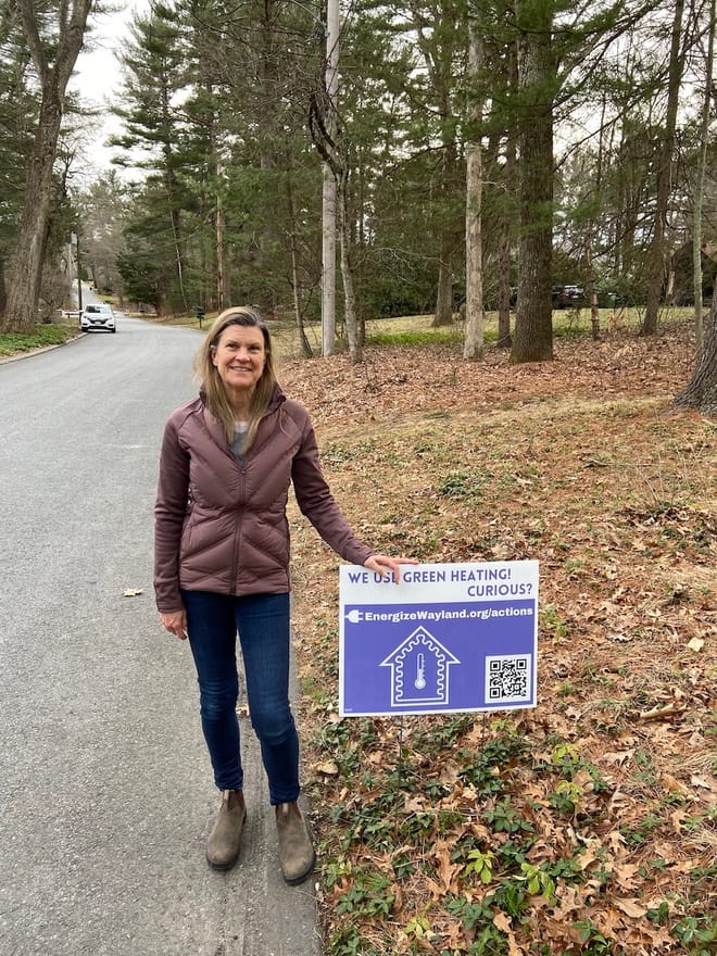 Woman with purple jacket stands in street next to yard sign with leaves on ground and trees in background.