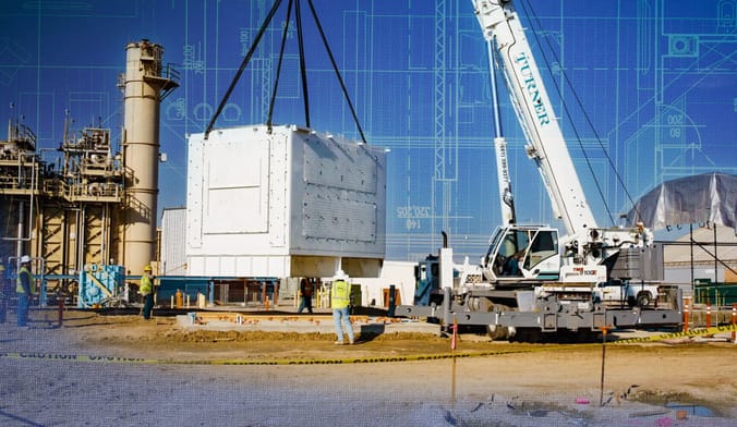 A crane lowers a large white metal box onto a construction site as workers in safety gear look on