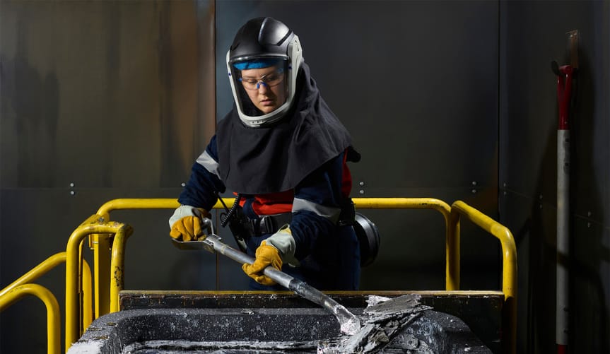 A worker wearing a safety mask and protective clothing shovels a silvery material in a black bucket