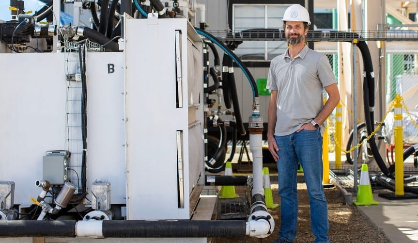 A man with a beard and a light skin tone wearing a white hard hat stands next to some industrial equipment