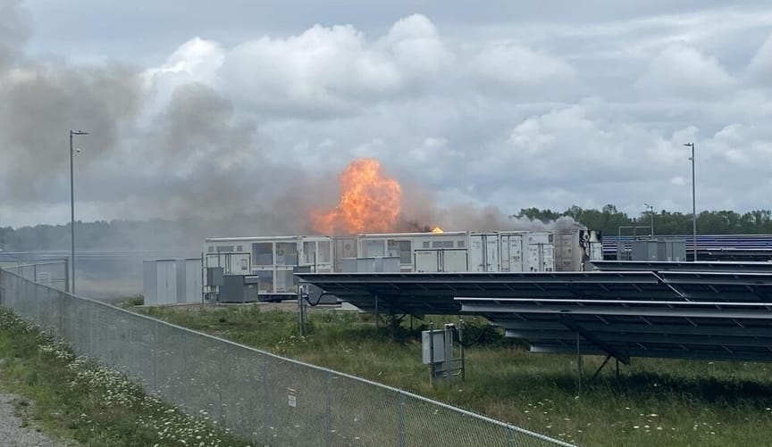 A large rectangular white metal container is on fire. It is next to solar panels and behind a fence.