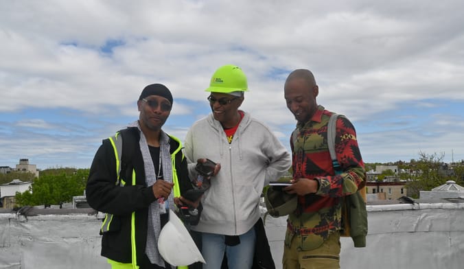 Three Black men stand on a roof, one wearing a neon green hardhat