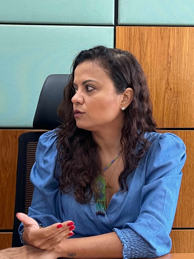 A women with long dark wavy hair wearing a blue top speaks and gestures while sitting in a conference room