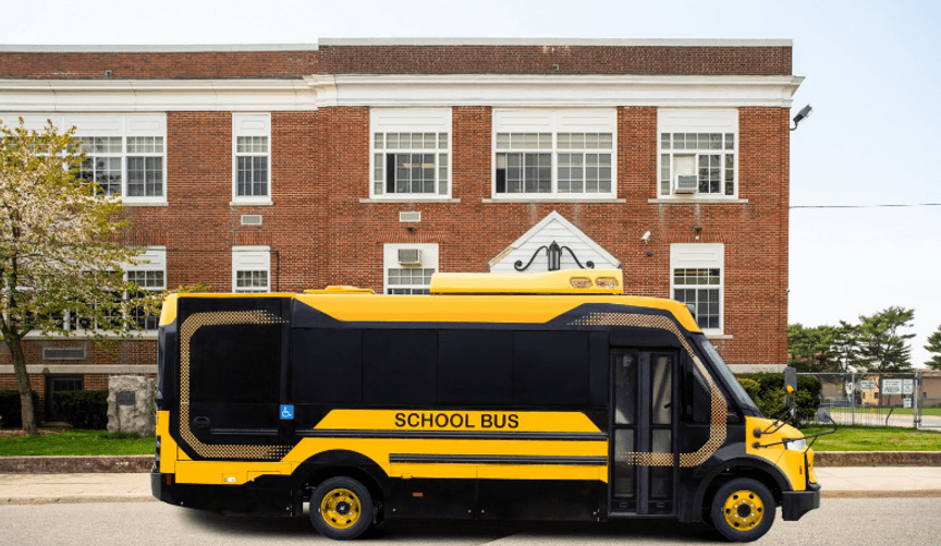 A sleek orange and black electric school bus is parked in front of a multistory red brick building.