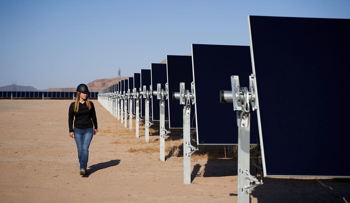 A woman wearing jeans and a black shirt walks alongside a large solar installation in the desert