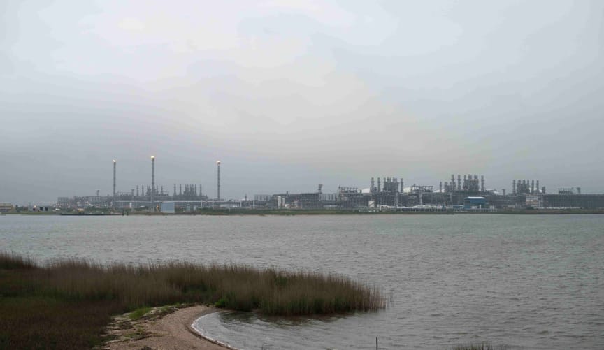 A very large industrial complex is seen on the distant shore of a lake. The sky is gloomy and gray