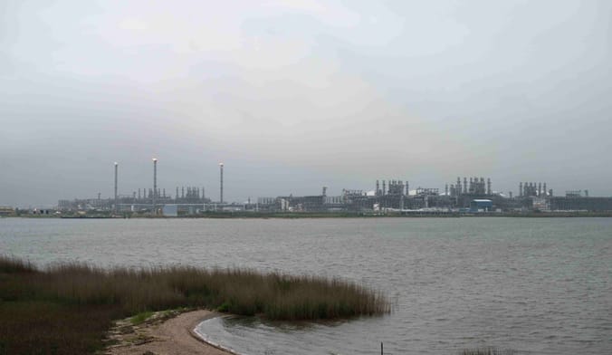 A very large industrial complex is seen on the distant shore of a lake. The sky is gloomy and gray