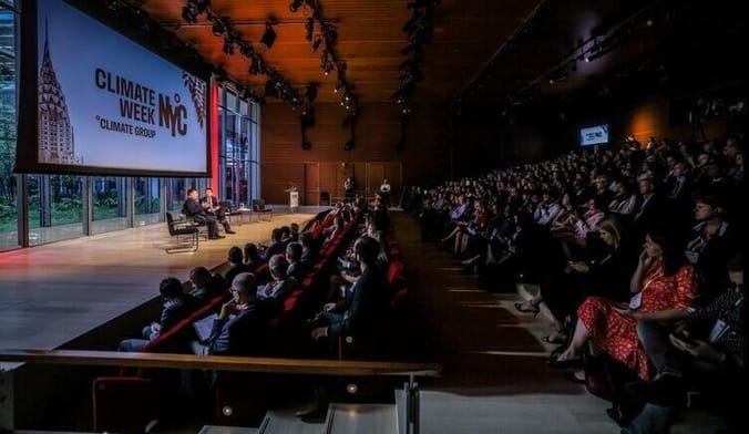 a large audience sits in an auditorium. Several people engage in a panel discussion onstage. A large sign says Climate Week
