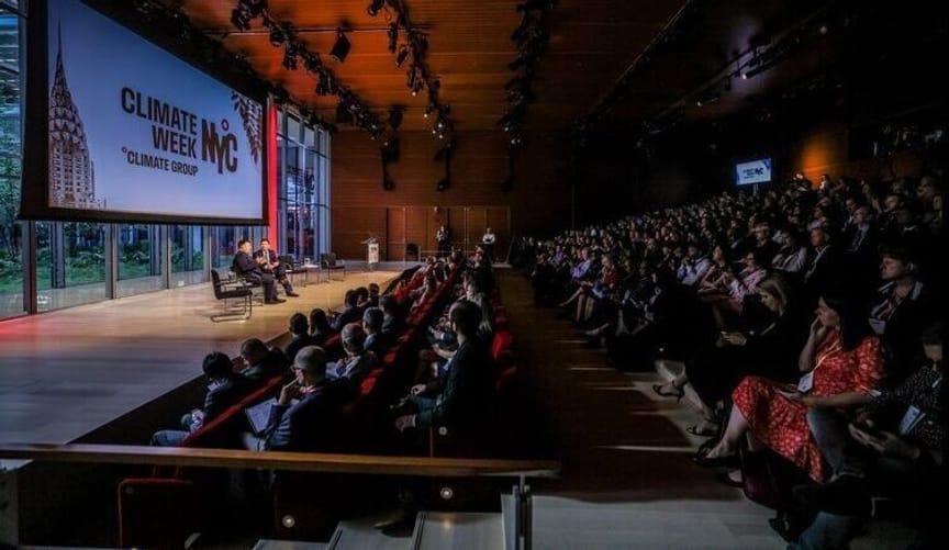 a large audience sits in an auditorium. Several people engage in a panel discussion onstage. A large sign says Climate Week