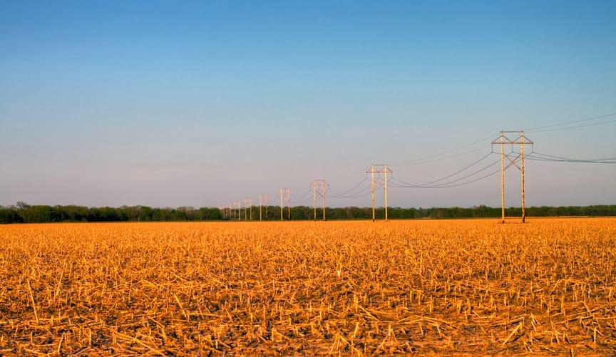 Rusty transmission lines stretch across the plains. In the foreground is a post-harvest cornfield with broken golden stalks