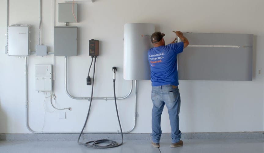A worker in a blue t-shirt and jeans installs a rectangular metal battery in a garage