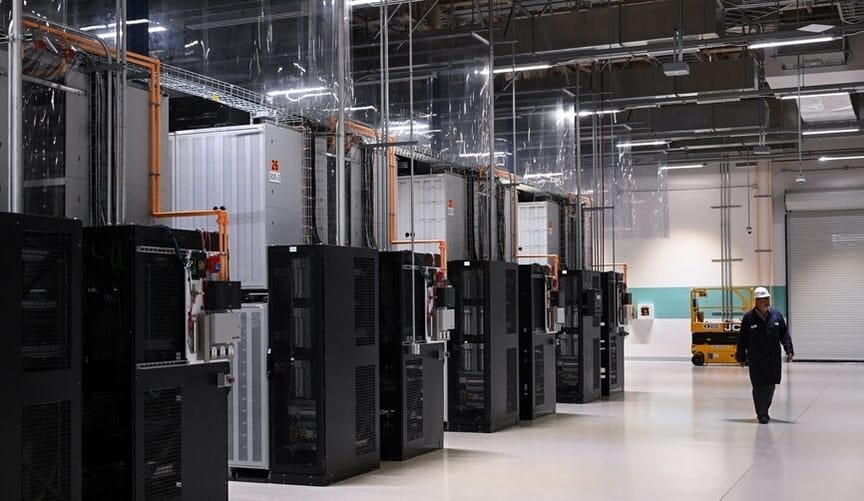 An employee in a hard hat and safety gear walks next to a row of large rectangular metal devices inside a warehouse