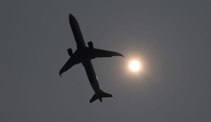 A passenger plane flies overhead in a gray sky next to a hazy sun
