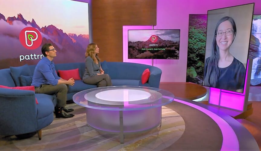 Two people sitting on a couch engage in conversation with a woman with long black hair and eyeglasses who is on video