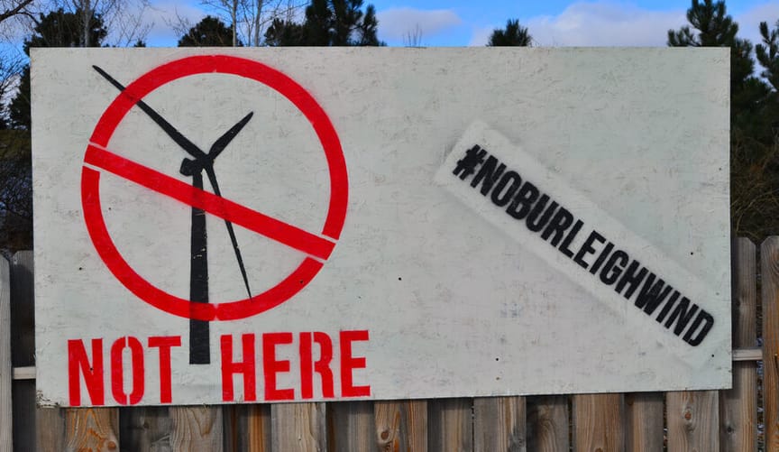 A sign affixed to a fence shows a wind turbine with a red slash across and the words "not here"