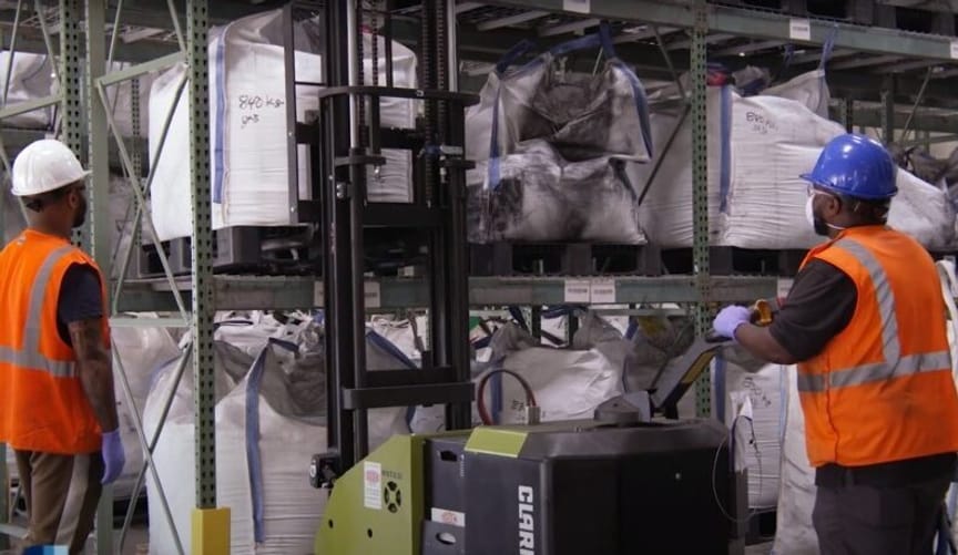 Two men in factory work gear look at a tall shelf piled with white bags