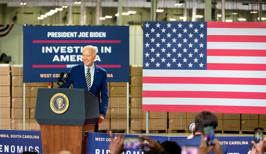 A white man with white hair wearing a blue jacket stands at a lectern in front of a crowd. Behind him is an American flag.