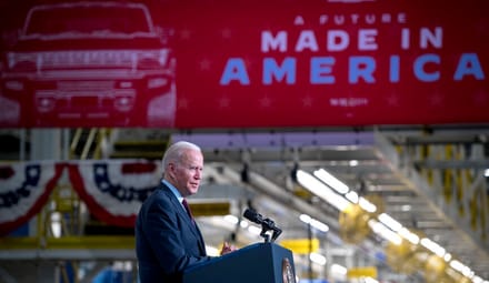 A man with white hair wearing a blue suit speaks at a lectern under a sign that reads "A future made in America"