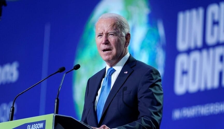 A man with white hair wearing a dark blue jacket and medium blue tie speaks into microphones at a podium