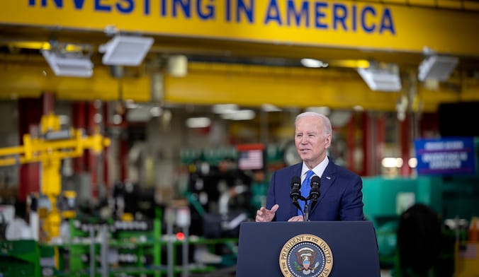 A man with light skin and white hair wearing a dark blue suit speaks at a presidential lectern on a factory floor