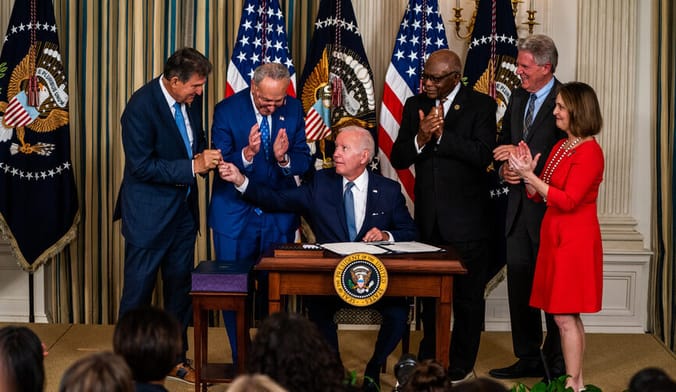 four men and one woman stand behind a desk where President Biden sits. US Flags and official insignia are in the background.