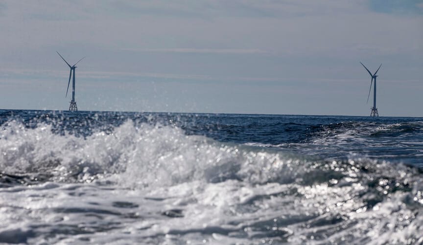 Two offshore wind turbines are seen in the distant background; in the foreground are choppy ocean waves and spray