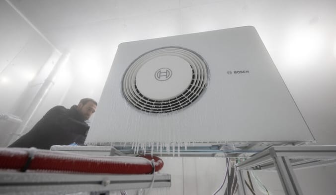 A man in dark clothing inspects a heat pump in factory facility. Icicles and frost are shown on the front of the heat pump.