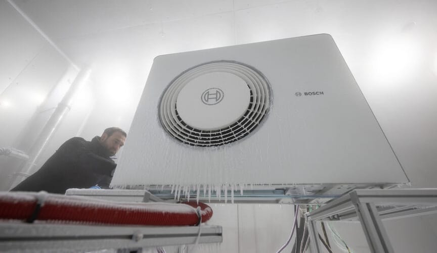 A man in dark clothing inspects a heat pump in factory facility. Icicles and frost are shown on the front of the heat pump.