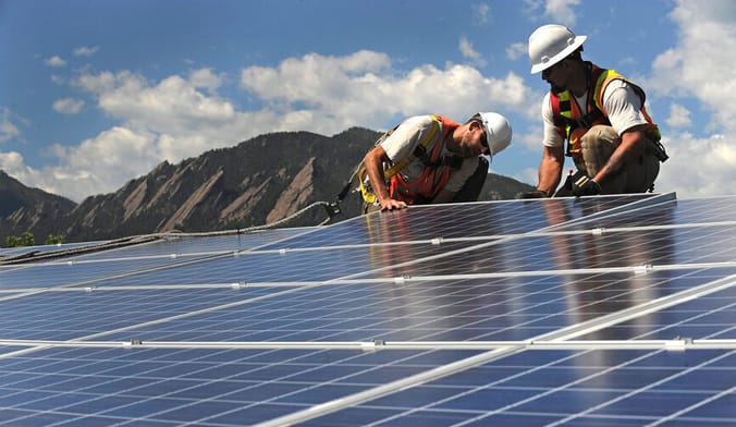Two men in white hard hats and safety vests slot a solar panel into place. A craggy mountain range is in the background.