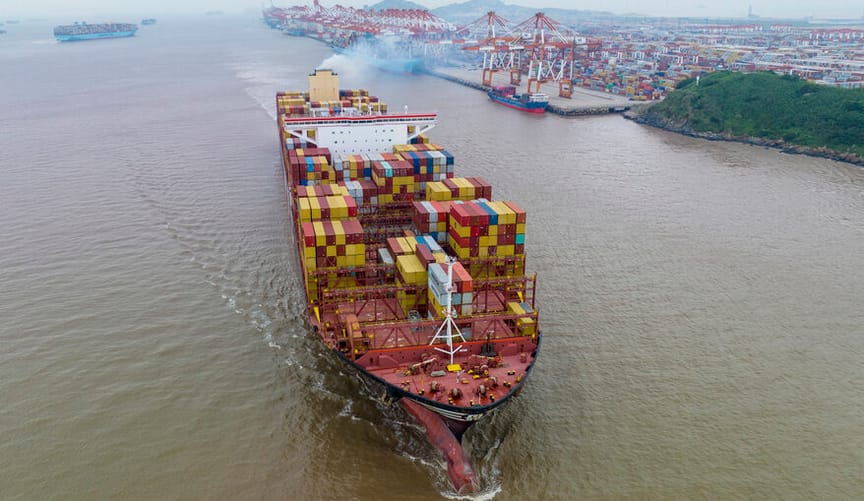 A large cargo ship piled high with colorful metal containers and spewing exhaust travels on the water
