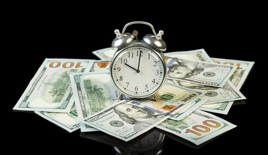 an old-fashioned alarm silver clock with bells on top sits on a pile of US $100 bills