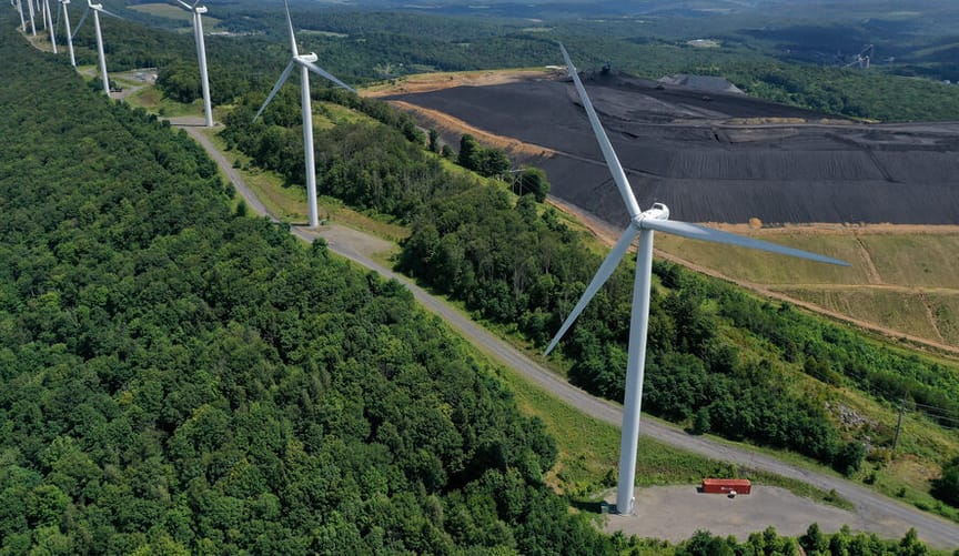 A row  of tall white wind turbines next to a coal mining operation amid a forested area