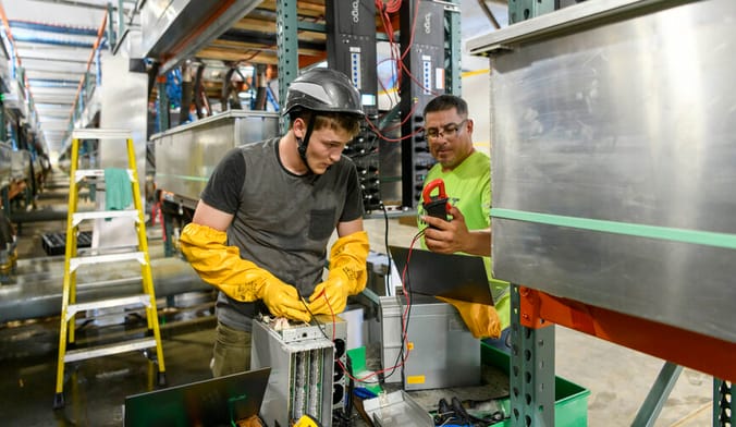 Two workers perform a service on mechanical equipment inside an industrial facility