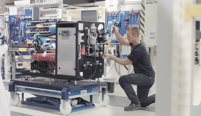A white man wearing black clothes kneels next to a mechanical device in a lab facility