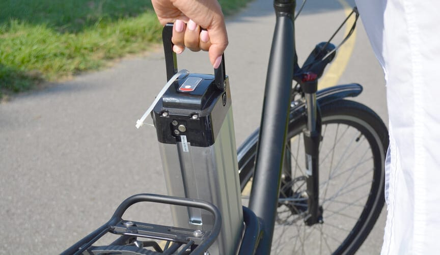 A hand with pink painted fingernails is seen removing a battery from an e-bike on an outdoor path