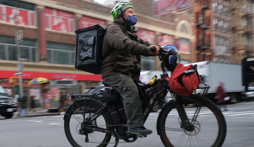 A man wearing a helmet, a mask and a winter coat rides an e-bike on a city street. He has a food-delivery backpack on.