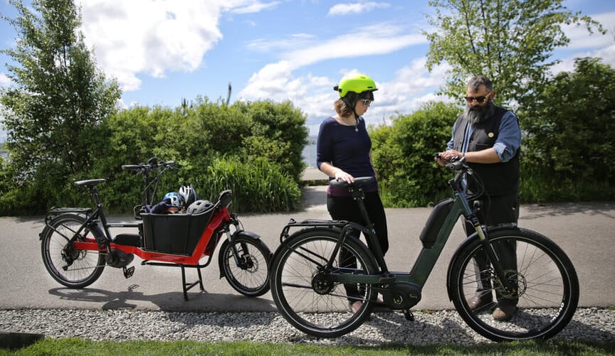 a woman in a neon green bike helmet stands next to an e-bike as a man in a vest and sunglasses explains its operation