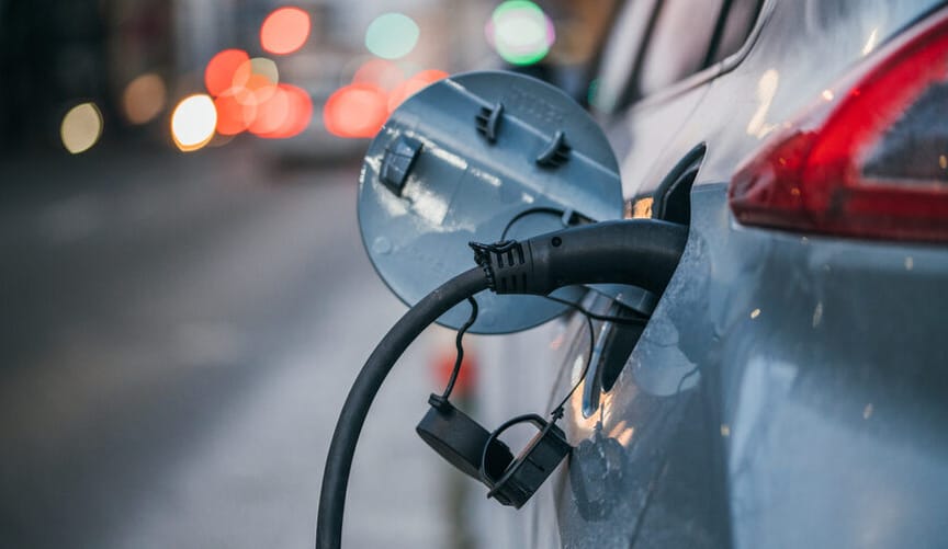 A blue car with an electric charger plugged into. Taillights blur in the background.